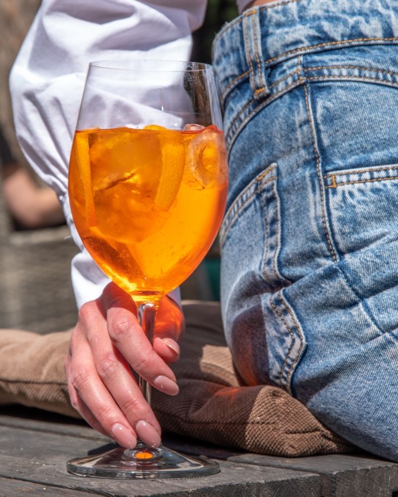 Person holding an orange cocktail in a glass with denim pants.