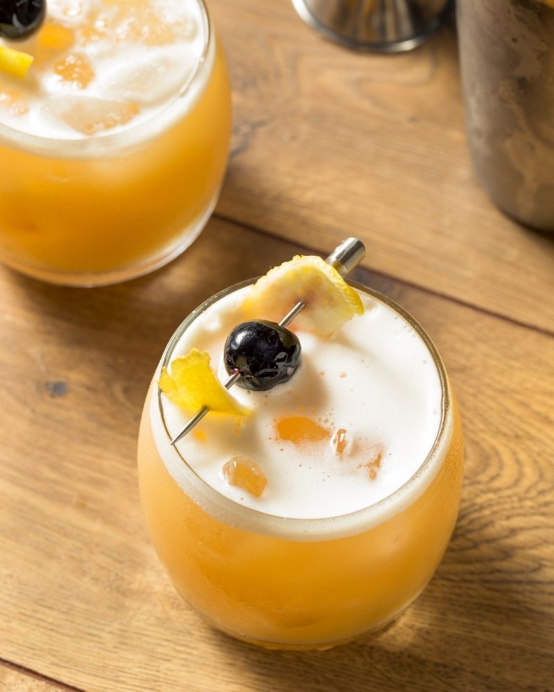 Two glasses of golden cocktails with foam a cherry, and a lemon wedge on a wooden surface.