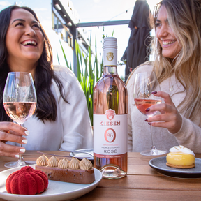 two women sitting at a picknic table socializing and enjoying glasses of giesen rose dealcoholized wine.  Table scape with bottle of Giesen 0% Rosé wine in center surrounded by delicious deserts.