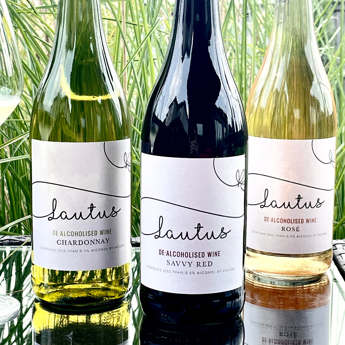 3 bottles of wine on a glass table outdoors with long tall grasses int he background.