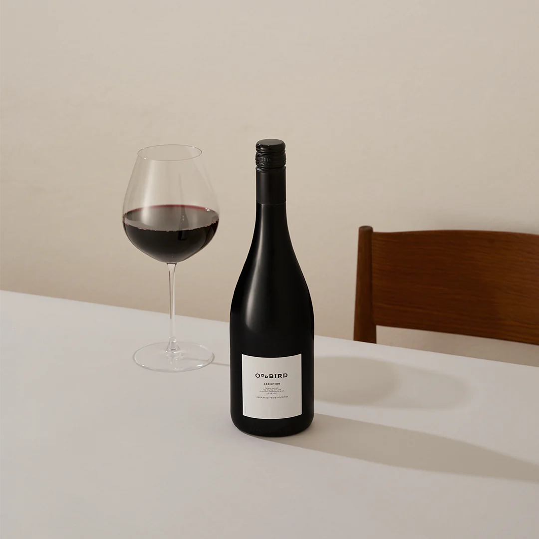 "Oddbird Addiction organic non-alcoholic red wine bottle and glass of red wine on a white table, set against a neutral background with a wooden chair — elegant table setting for premium dealcoholized Tempranillo."