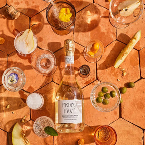 This image features a beautifully styled flat lay of Prima Pave Blanc de Blancs non-alcoholic sparkling wine. The bottle is surrounded by an elegant assortment of glasses, fresh fruits, and garnishes, emphasizing a sophisticated and celebratory vibe.