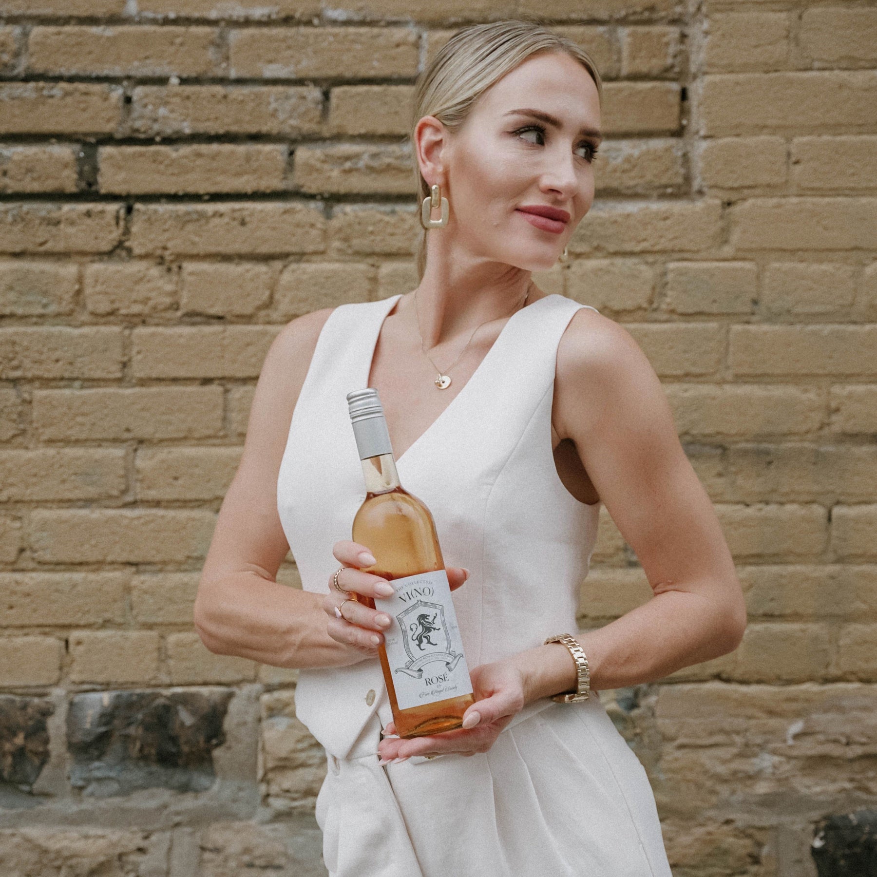 A woman stands against a brick wall, holding a bottle of rosé wine labeled "Vino Collection." She is dressed in an elegant, light-colored sleeveless outfit and wears gold hoop earrings, a necklace, and a watch, looking to the side with a slight smile. The setting is casual yet refined, complementing the sophisticated feel of the wine she holds.