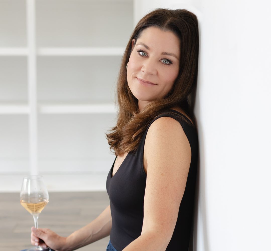 Woman sitting against a wall holding a glass of white wine against a neutral background
