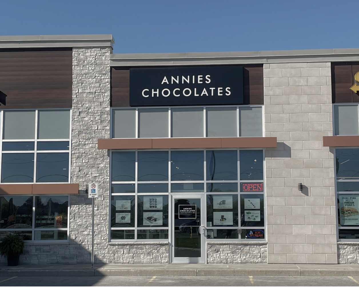 Front exterior of a commercial building with a large black sign reading 'ANNIES CHOCOLATES' above the entrance. The facade combines gray stone veneer and dark wood panels, with large glass windows and doors.