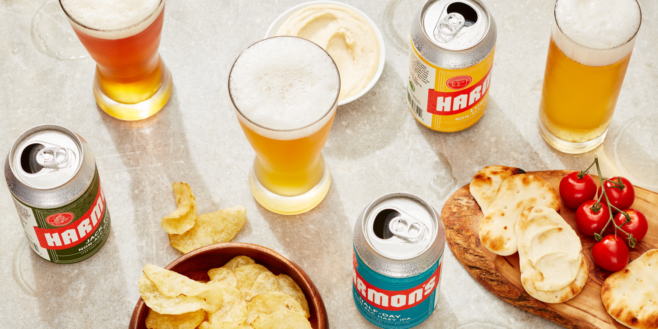 The image shows an assortment of non-alcoholic beers by Harmon’s, displayed alongside snacks on a light-colored surface. There are three cans of Harmon’s beverages in different varieties, with labels in green, blue, and yellow, and each can is partially opened. Several glasses of beer are poured with foamy tops. The snacks include a bowl of potato chips, a small wooden board with mini naan breads spread with hummus, and a bunch of fresh cherry tomatoes on the vine. The scene has a casual, inviting feel, per