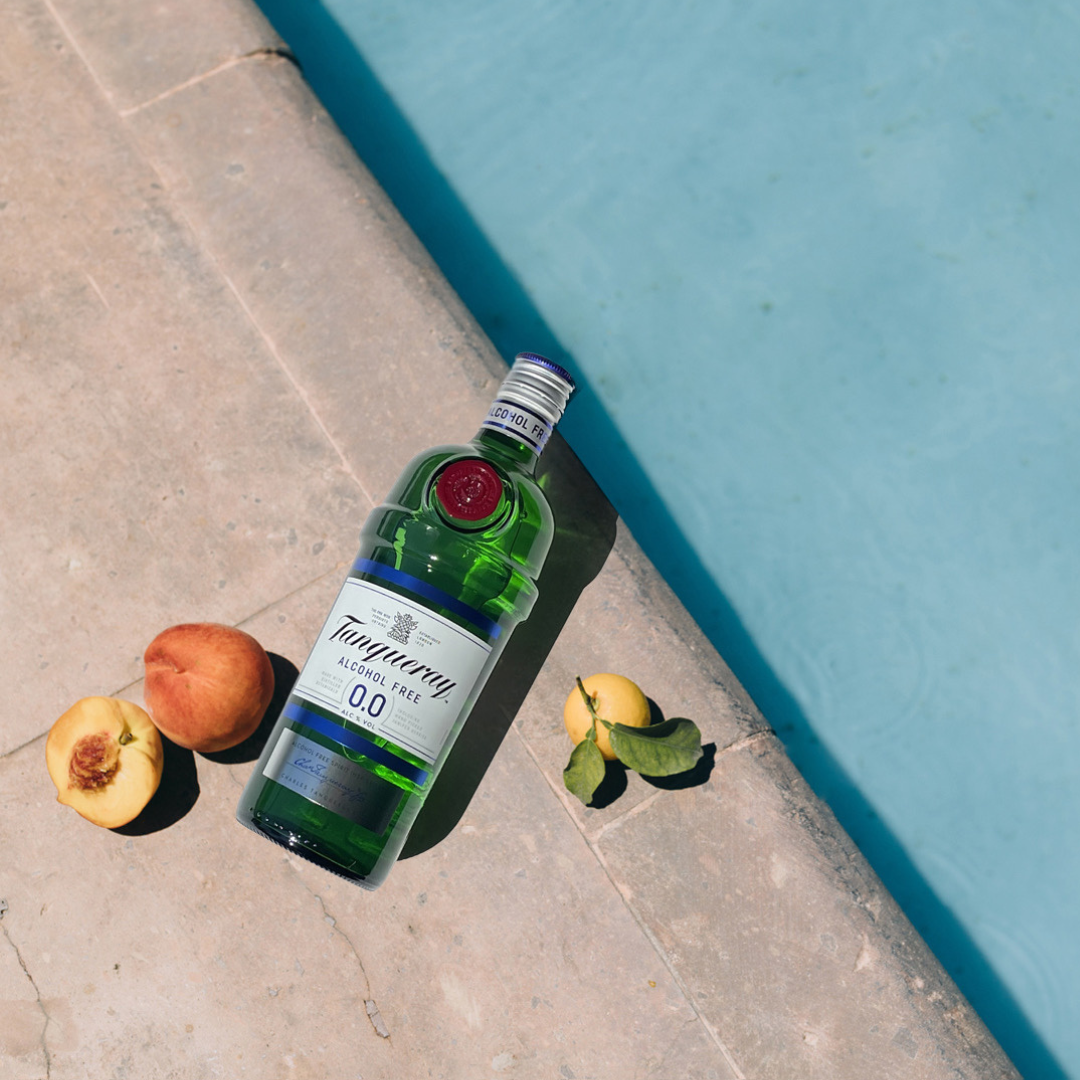 A bottle of Tanqueray alcohol free gin laying flat on concrete pool side with peaches and fruit displayed beside the bottle.  The label reads Tanqueray 0.0% and is green with white labels and blue and red accent colours.