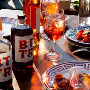A vibrant summer table scene featuring BTR Non-Alcoholic Spirit bottles paired with colorful aperitif-style drinks in elegant glasses. Surrounding the drinks are plates of fresh Mediterranean-inspired dishes, including tomatoes, cured meats, and mozzarella, all basking in warm sunlight, evoking a lively, social gathering.