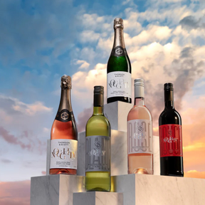 Five bottles of Noughty non-alcoholic wines displayed on marble pedestals against a backdrop of a dramatic, colorful sky. The lineup includes sparkling rosé, sparkling chardonnay, white wine, still rosé, and red wine, showcasing the variety of alcohol-free options from Noughty.