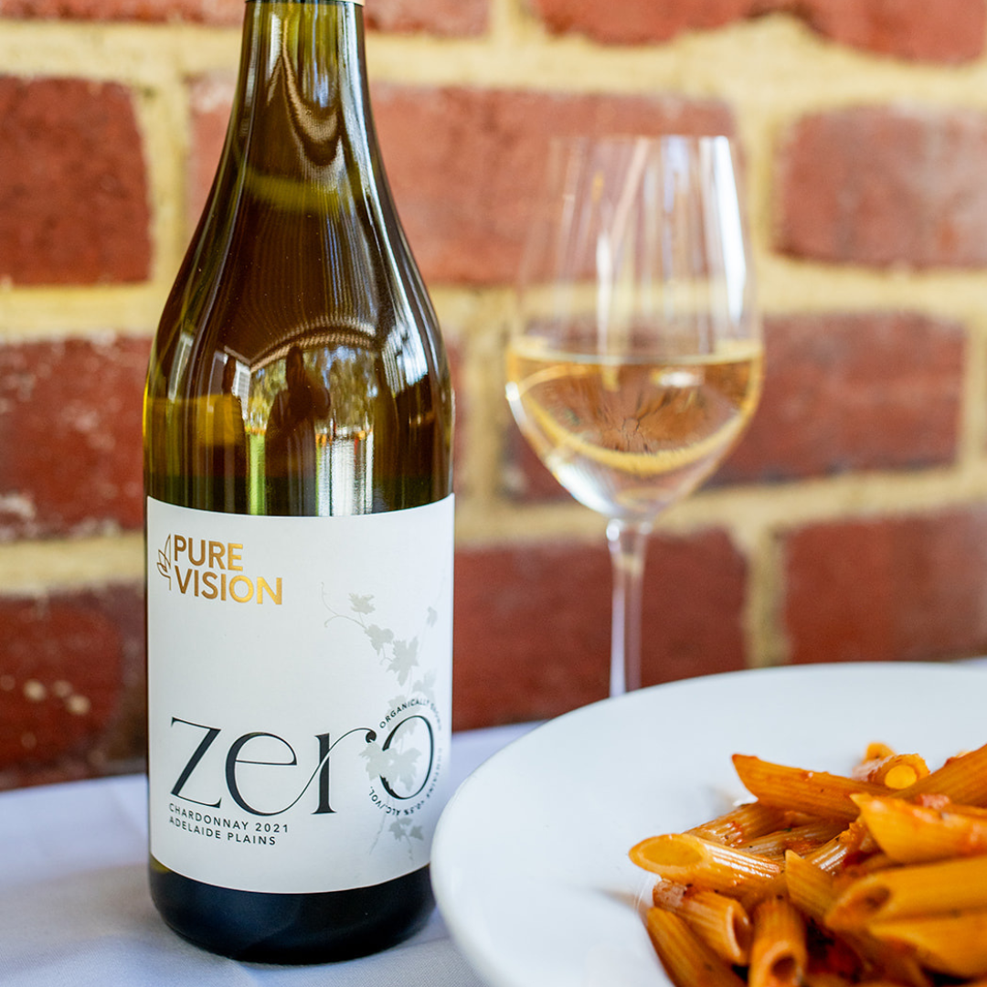 A bottle of Pure Vision Zero Non-alcoholic chardonnay wine on a table set with a glass of white wine and a plate of pasta with red sauce.  Backdrop is a red brick wall of the tratoria.