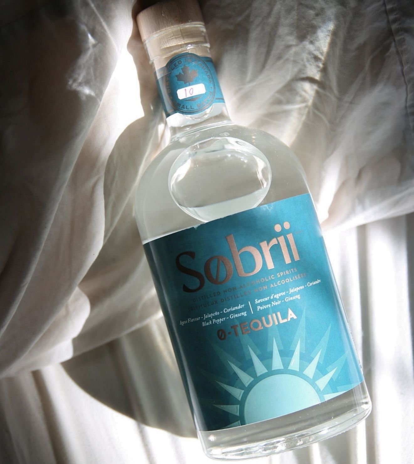 Bottle of Sobri-0 Tequila with a blue label with a bright sun on a white textured background that looks like wrinkled sheets.  Hard sunlight shadows reflecting on bottle creating shadows. 
