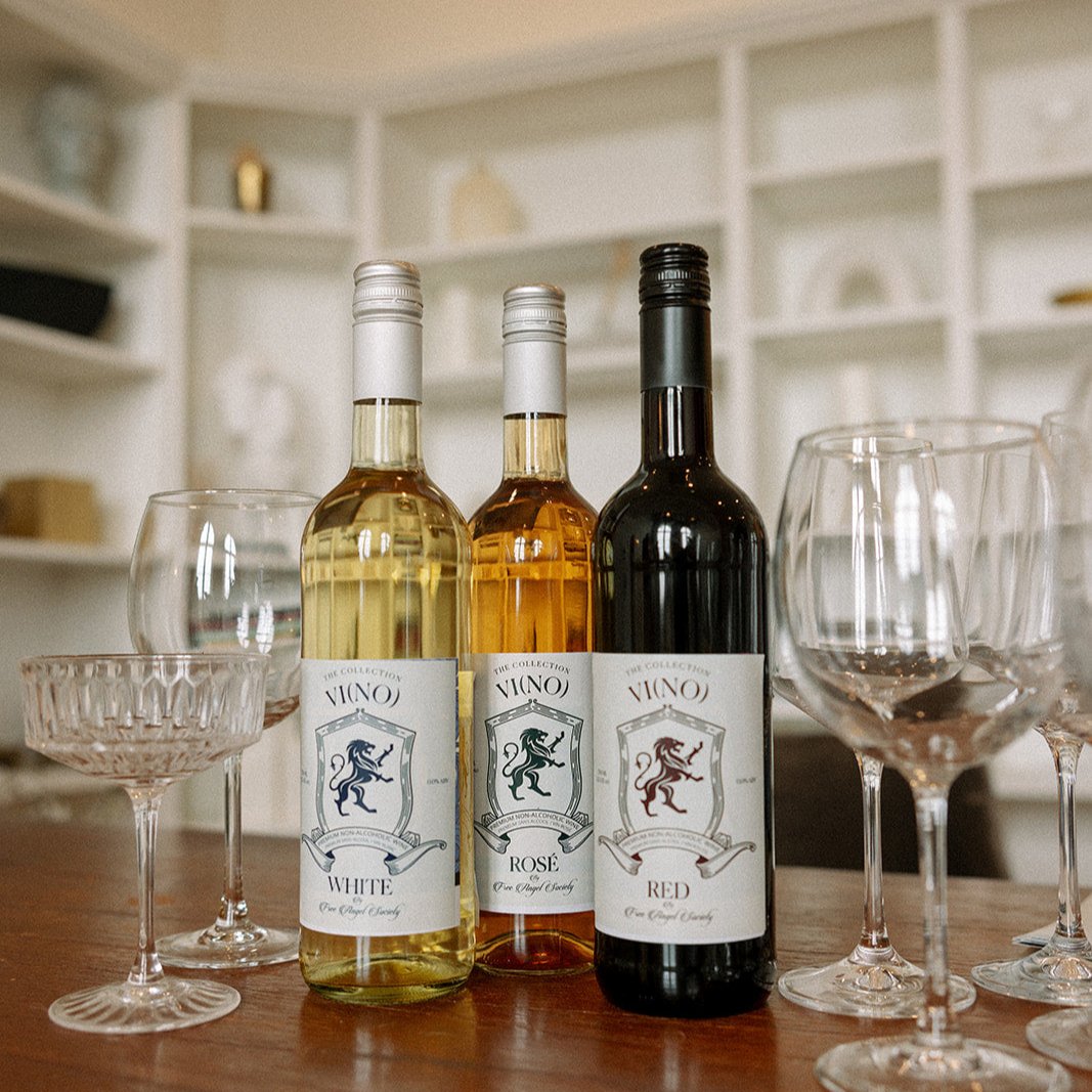 Three bottles of wine, labeled as "Vino The Collection" in white, rosé, and red varieties, are displayed on a wooden table. The bottles are surrounded by an assortment of empty wine glasses, including stemmed glasses and one coupe glass. In the background, there is a softly lit, modern shelving unit with decorative items, adding to the elegant and inviting atmosphere.