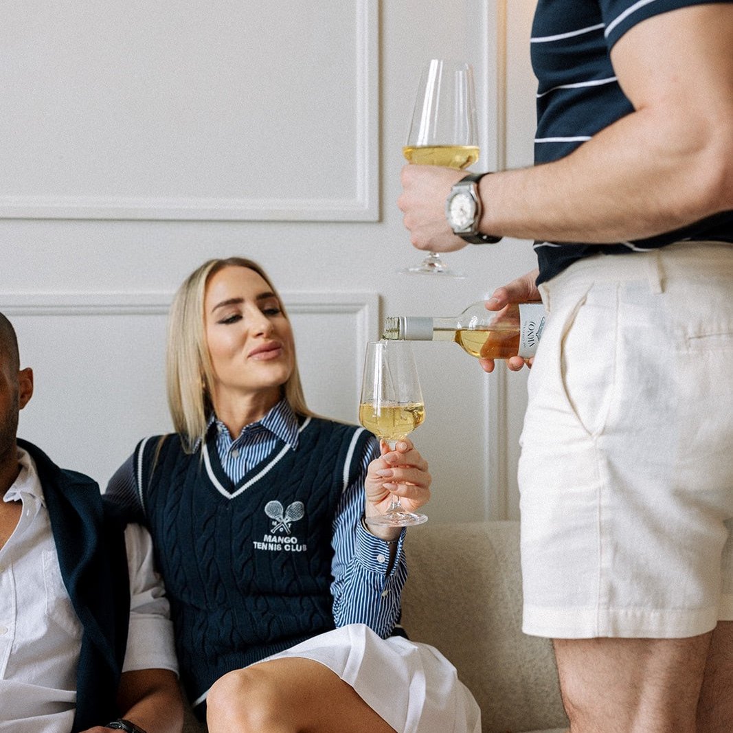 A casually dressed group of friends enjoy a relaxed gathering indoors. A man in a striped shirt and white shorts pours white wine into a glass held by a woman seated on a couch. She is wearing a navy blue tennis club vest over a striped shirt and smiling as she receives the pour. Another man, partially visible, sits beside her in casual summer attire, creating a warm and sociable atmosphere.