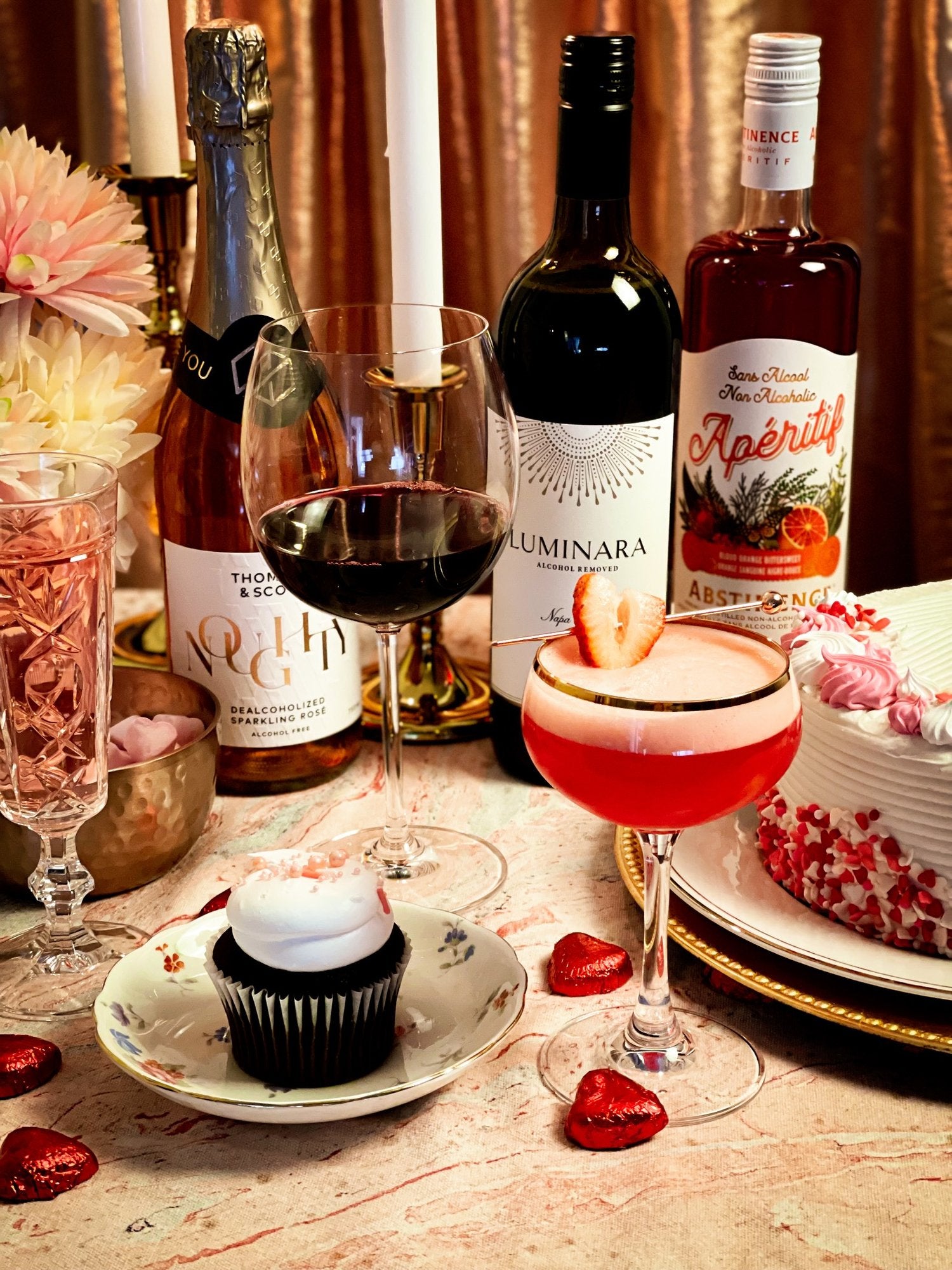 group shot of luminara red wine in dark bottle, abstinence aperitif red spirit with organges on label, bottle of noughty sparkling rose wine, glass of red wine and delicious red cocktail with strawberry garnish, on a table scarpe with a valentine cake, chocolate cupcake and heart shaped chocolates.  flowers and candles in background